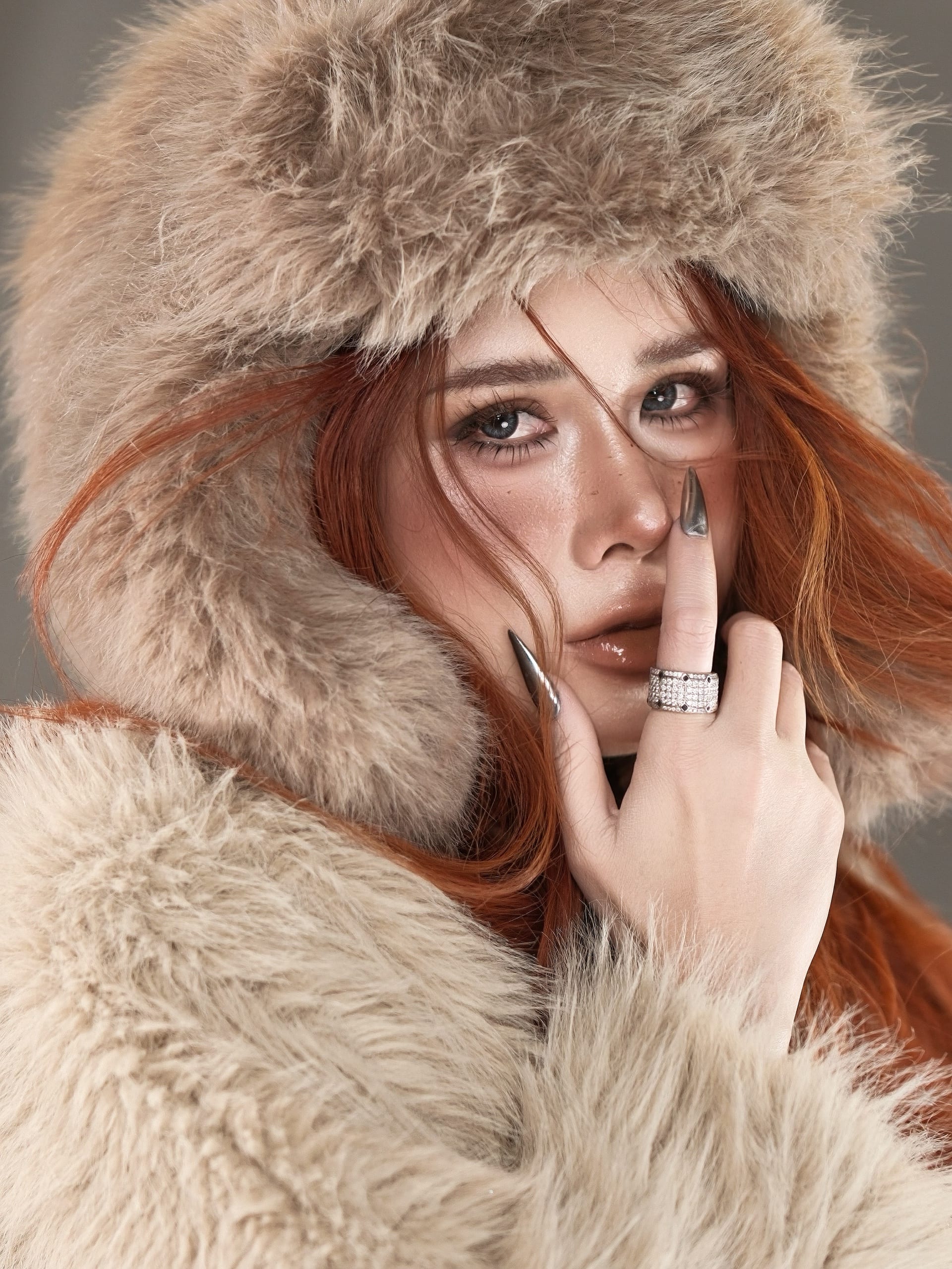 Chân dung beauty: tóc đỏ đồng, mắt kẻ khói nâu, da căng bóng giữ tàn nhang, tay đưa lên môi; mũ và áo lông màu be.
