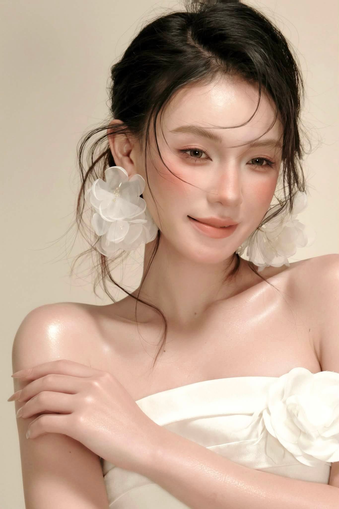 Chân dung nền sáng: nền da căng bóng, mắt nâu ấm, môi cam đất; hoa vải trắng lớn làm khuyên tai, áo quây trắng.